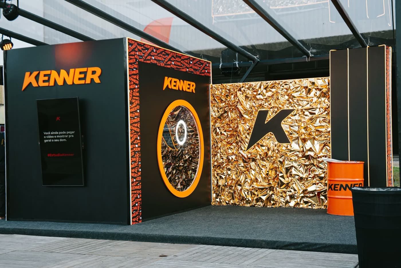 展台外立面：品牌标识、贴着金色纸并带有字母 K 的墙面，以及可以看进录音亭内部的圆形视窗