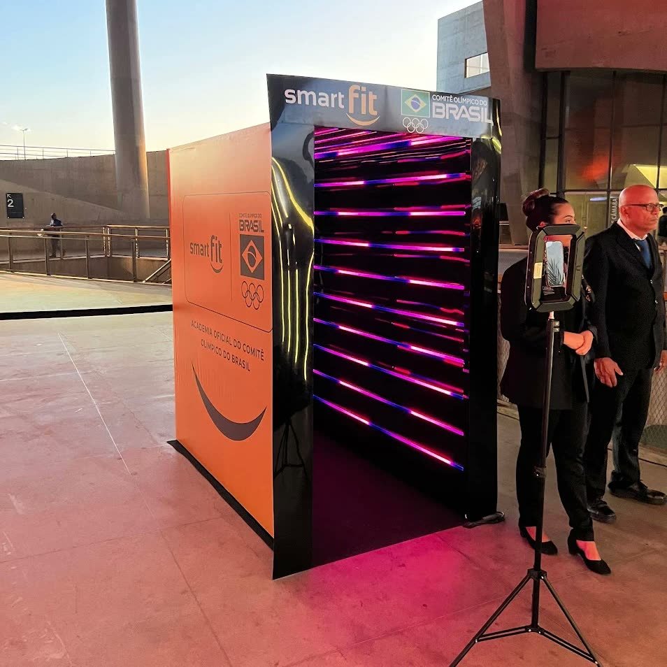 Túnel de LED visto de lado, com as fitas de luz rosa e azul por dentro e a lateral laranja com a marca